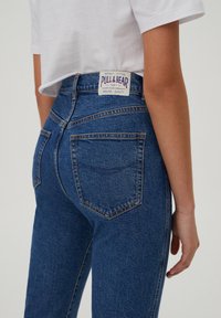 Personne portant un jean en denim bleu taille haute avec une étiquette de la marque Pull & Bear et un T-shirt court blanc sur un fond uni.