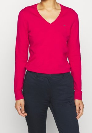 Pull à col en V, à manches longues, de couleur rose vif, fabriqué en matériau doux. Petit logo bleu et blanc sur la poitrine. Associé à un pantalon foncé.