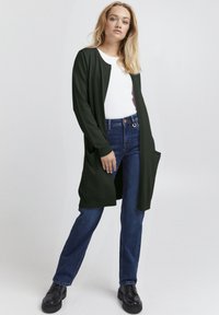Donkergroen cardigan met lange mouwen, open voorkant en zijzakken, gecombineerd met een witte blouse en blauwe spijkerbroek. Platte zwarte schoenen.