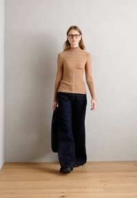 Bežinė banguota aukštakaklė palaidinė, derinama su plačiomis tamsiai mėlynomis džinsų kelnėmis. Modelis laiko juodą rankinę, eidamas prieš neutralų pilką foną.