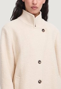 Cremefarvet fleecejakke med høj krave, med knaplukning og sidelommer. Blød tekstur og minimalistisk design.
