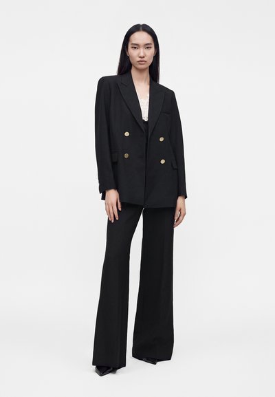 Femme debout portant un blazer noir croisé avec des boutons dorés, un pantalon noir à jambes larges et un top en dentelle blanche en dessous.