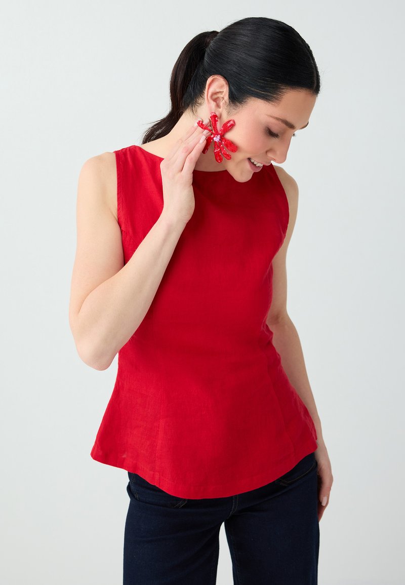 Femme aux cheveux noirs portant un haut rouge sans manches touchant une grande boucle d'oreille fleur rouge, posant sur un fond gris clair.