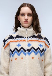 Fleece zip-up jakke i creme, med et geometrisk mønster med orange, blå og sorte accenter. Blød tekstur, høj kravedesign.