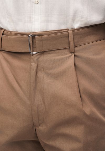 Κοντινό ανέκφραστο της μέσης με μπεζ παντελόνια με ζώνη από τον ίδιο ύφασμα και ασημί ορθογώνια αγκράφα, σε συνδυασμό με λευκό καρό πουκάμισο κρυμμένο μέσα.