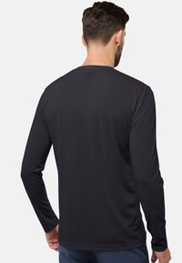 Mann mit kurzem braunem Haar, der ein schlichtes schwarzes Langarmshirt und blaue Hosen trägt, von hinten vor einem weißen Hintergrund gesehen.