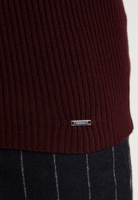 Maglione a coste bordeaux con orlo decorato da un'etichetta argentata. Il tessuto grigio scuro sottostante presenta righe bianche. Texture morbida.
