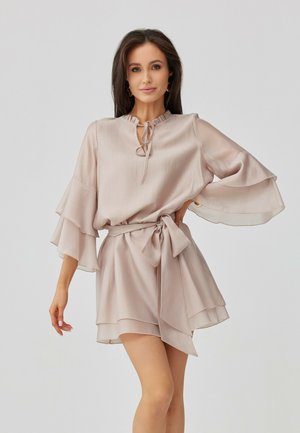 Žena s dlouhými tmavými vlasy, oblečená v lehkých béžových vrstvených šatech s širokými zvonovými rukávy a páskem v pase, stojící před šedým pozadím.