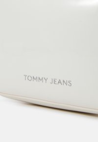 Bílá kabelka s hladkým, lesklým povrchem, se vyraženým logem "TOMMY JEANS" v stříbrné barvě na přední straně. Žádné další kovové doplňky nejsou viditelné.