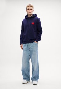 Marineblå hettejakke med rød logo-patch, brukt sammen med lyseblå vide jeans og hvite sneakers. Glatt stoff, uformell design.