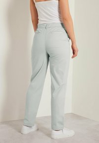 Pantaloni verde chiaro realizzati in tessuto morbido, dotati di tasche posteriori e vestibilità comoda, abbinati a sneaker bianche su un pavimento grigio.