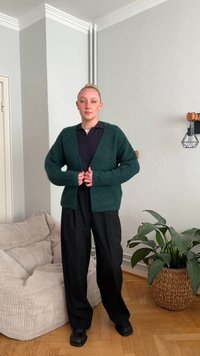 Ciemnozielony sweter na drutach o luźnym kroju, z otwartym przodem i długimi rękawami, zestawiony z czarnymi szerokimi spodniami i czarnymi butami.