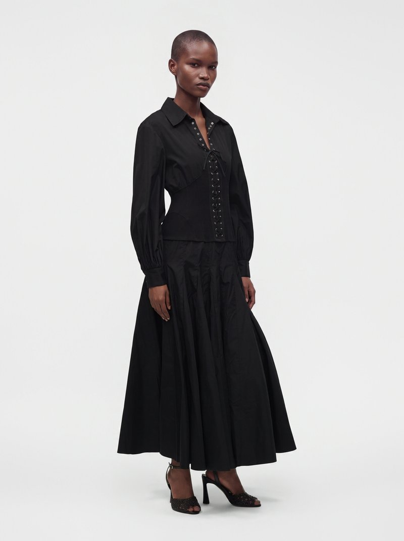 Femme portant une longue jupe noire et un chemisier noir avec un détail à lacets devant, debout devant un fond clair uni.