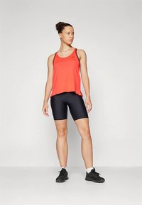 Femeie în picioare purtând un top sportiv roșu fără mâneci, pantaloni scurți negri de ciclism, șosete albe și adidași negri, pe un fundal simplu deschis.