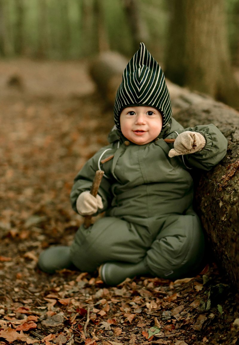 Tuta imbottita verde con cappuccio e guanti beige. Il cappuccio presenta un motivo a righe. Bambino seduto su un tronco in un ambiente forestale.