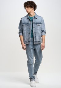 Chaqueta de denim azul claro con diseño clásico, combinada con una camisa verde azulado y jeans azul claro. Zapatillas blancas completan el atuendo.