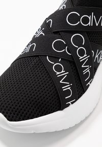 Černá sportovní bota s texturovaným síťovaným svrškem a výraznými bílými popruhy s logem Calvin Klein. Podrážka je bílá a poskytuje pohodlné odpružení.
