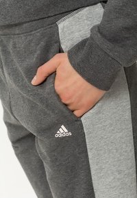 Main reposant dans la poche d'un pantalon de survêtement Adidas gris foncé et clair avec le logo Adidas visible sur la jambe.