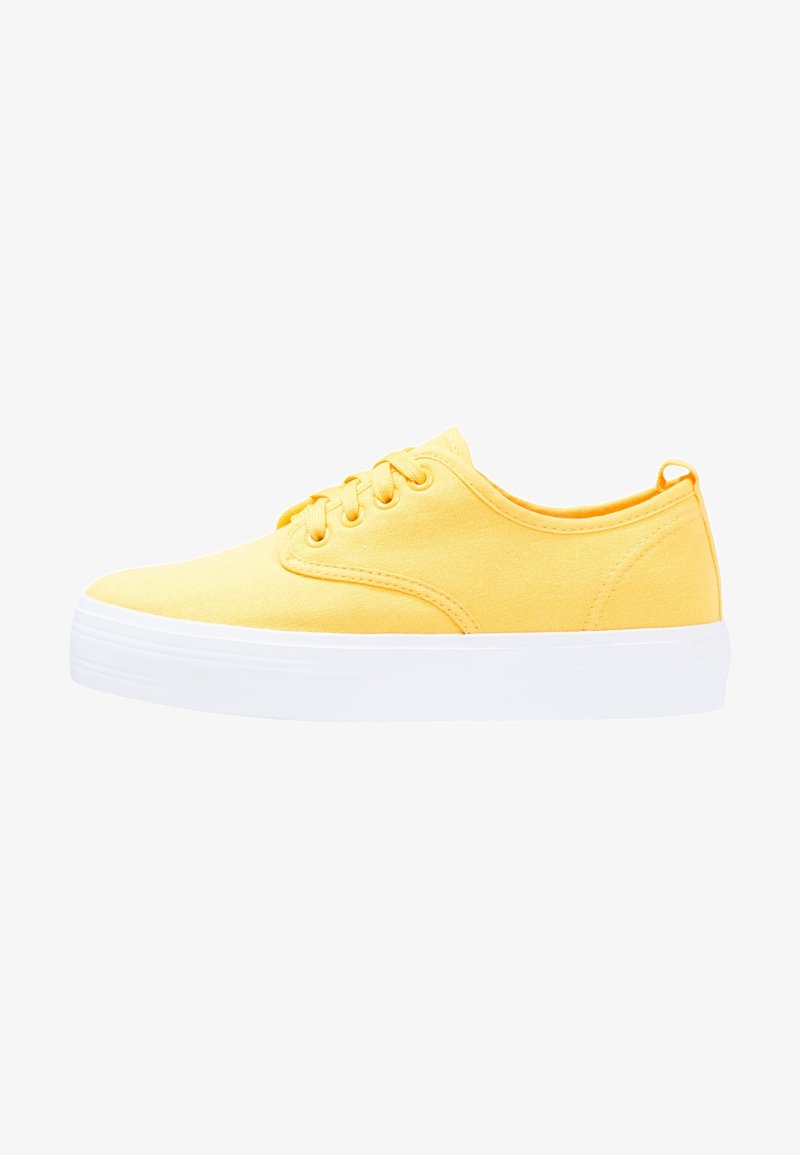 Des baskets en toile jaunes avec des semelles en caoutchouc blanches, à lacets, à bout arrondi et à texture lisse. Style minimaliste sans motifs visibles.