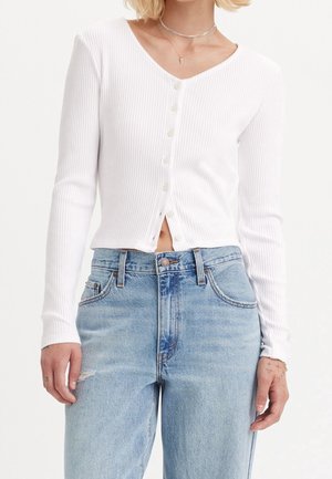 Vrouw draagt een wit geribd lang mouwen knooptopje en hooggetailleerde lichtblauwe jeans, met eenvoudige zilveren kettingen zichtbaar.