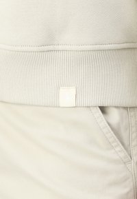 Hellebeige Sweatshirt mit geripptem Saum und Bündchen, mit einem kleinen weißen Stoffetikett, auf dem "CK" auf der Seite angenäht ist.