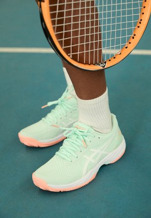 Leg in white sock wearing mint green and peach tennis shoes standing on blue court holding orange tennis racquet.