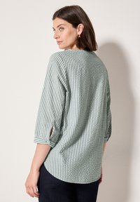Blusa a righe verde e bianca con maniche a tre quarti, tessuto testurizzato, orlo arrotondato e dettaglio con bottoni sui polsini delle maniche.