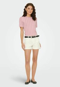 Maglietta lavorata a maglia di un delicato rosa chiaro con design a coste, abbinata a pantaloni corti bianchi e una cintura nera con borchie. Ballerine nere completano l'outfit.