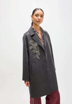 Mujer con cabello oscuro recogido en una coleta lleva un abrigo gris con un adorno floral sobre una camisa estampada y pantalones granate.