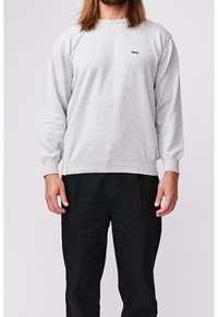 Homme portant un pull léger à manches longues gris clair avec un petit logo noir "Makia" et un pantalon noir, debout devant un fond uni.