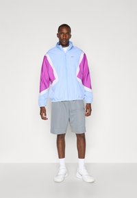 Nike windbreaker in light blue with purple and white sections, featuring a zip front, elastic cuffs, and a casual fit, paired with gray shorts.