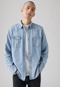 Lichtblauw denimhemd met een knoopsluiting, borstzakken en contrasterende stiksels, gedragen over een wit ondershirt.
