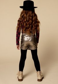 Kind met lang golvend haar, gekleed in een zwarte hoed met brede rand, een langärmige top met hartpatroon, metallic shorts, zwarte maillots en beige laarzen, van achteren gezien.