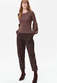 Vrouw met lang haar die een aansluitende, geribbelde bruine trui met pofmouwen draagt, donkerbruine broek met knopen bij de enkels en bruine laarzen met hak.