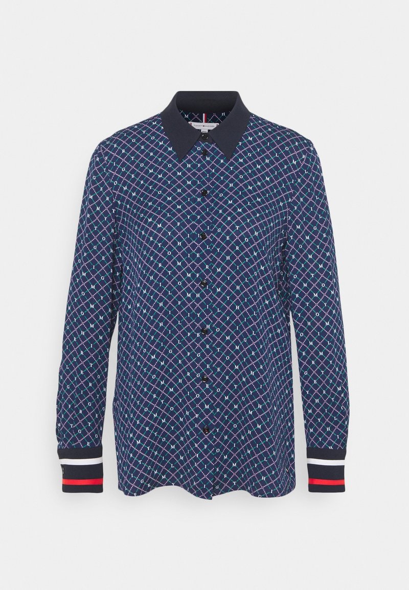 Chemise à manches longues bleu marine avec un motif géométrique blanc et rouge, col noir et poignets rayés noir, blanc et rouge.