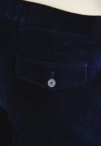 Ciemnoniebieskie spodnie z korduroy z tylną kieszenią, ozdobioną okrągłym srebrnym guzikiem. Tekstura jest prążkowana, co wskazuje na miękki i wytrzymały materiał.