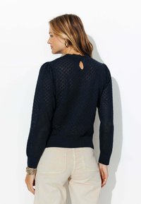 Pull bleu marine avec motif en losange, tricot texturé, encolure ronde, manches longues bouffantes et détail en forme de clé à l'arrière, associé à un pantalon clair.