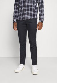 Man wearing navy slim-fit pants with a navy, white, and orange plaid shirt and white sneakers, standing against a plain background.