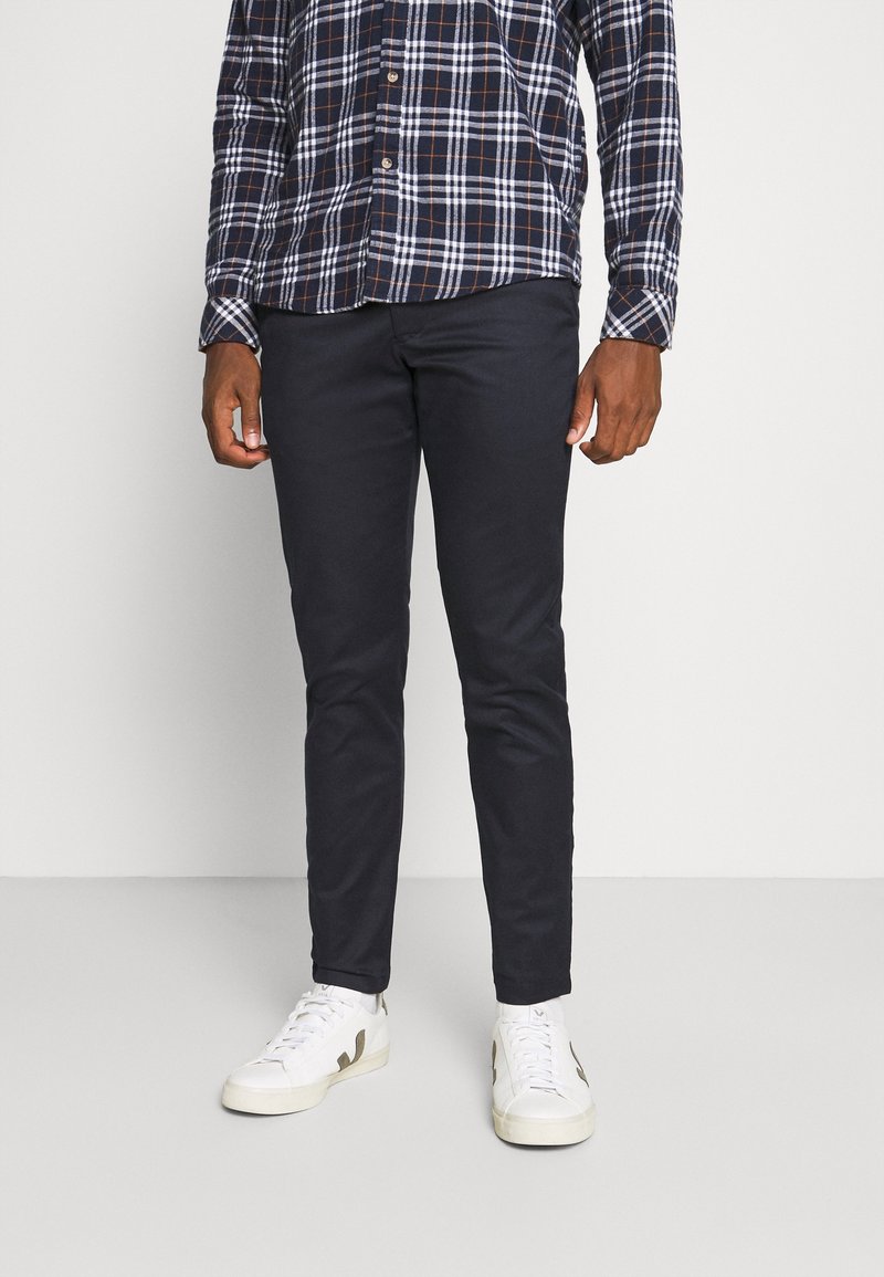 Man wearing navy slim-fit pants with a navy, white, and orange plaid shirt and white sneakers, standing against a plain background.