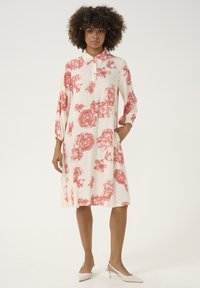 Rochie florală cu imprimeuri de flori roșii pe un fundal crem, guler scurt, mâneci lungi și buzunare laterale. Purtați cu pantofi slingback albi.