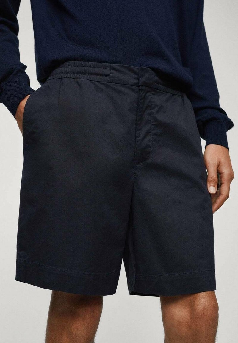 Personne portant un short noir à taille élastique et un haut à manches longues foncé, les mains posées près des poches, debout devant un fond uni.