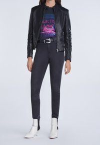 Chaqueta de moto de cuero negro con acentos ribeteados en los hombros, combinada con una camiseta gráfica de colores neón, jeans ajustados oscuros y botas de tobillo blancas.