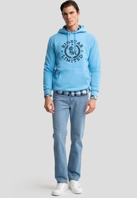 Lichtblauwe hoodie met capuchon en een zwart logo, gecombineerd met lichte denim jeans en witte sneakers.