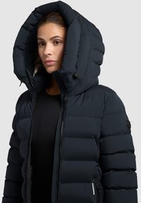 Chaqueta acolchada azul marino con una gran capucha, diseño de acolchado, cierre frontal con cremallera y parche de logo en la manga. Tejido suave con una textura lisa.