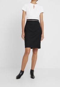 Bílý top s detaily ve tvaru klíčové dírky v kombinaci s černou tužkovou sukní. Obsahuje kontrastní pás. Modelka má na sobě černé kotníkové boty.