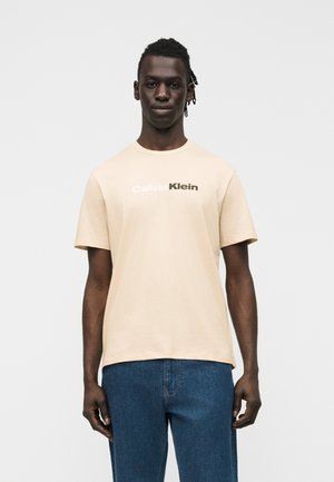 Tumeaga nahaga ja palmikutega mees kannab beežikat Calvin Kleini t-särki ja siniseid teksaseid, seistes ühetaolise hele tausta ees.