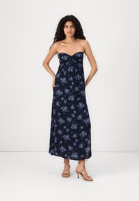 Navy floral maxi dress with a strapless design, featuring a knotted front detail. Light fabric flows to the floor. Tan strappy heels.