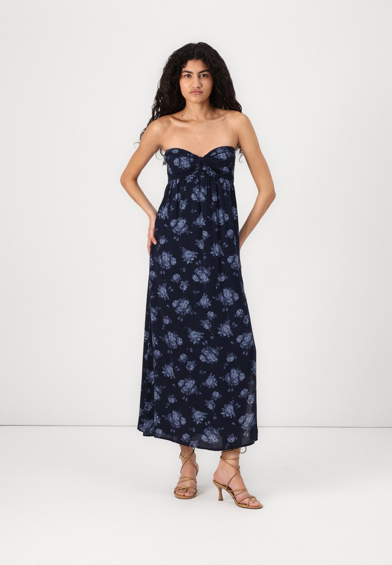 Navy-blaues, blumiges Maxi-Kleid mit trägerlosem Design und geknotetem Vorderteil. Leichter Stoff, der bis zum Boden fließt. Tanfarbene, geschnürte High Heels.