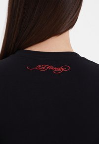 Black cotton t-shirt featuring a red embroidered "Ed Hardy" logo on the back near the neckline. Smooth texture, crew neckline design.