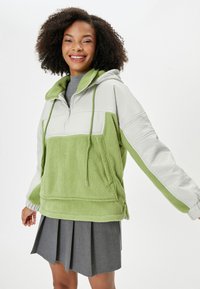 Sudadera de forro polar verde y beige con capucha, media cremallera y cordones. Presenta una superficie texturizada y un ajuste relajado.